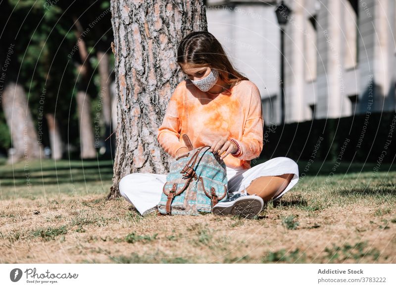 Junge Frau in Maske schaut in Tasche in der Nähe von Baum Schüler sitzen stöbern Rasen Universität Mundschutz Pandemie Madrid Spanien Suche Hochschule Bund 19
