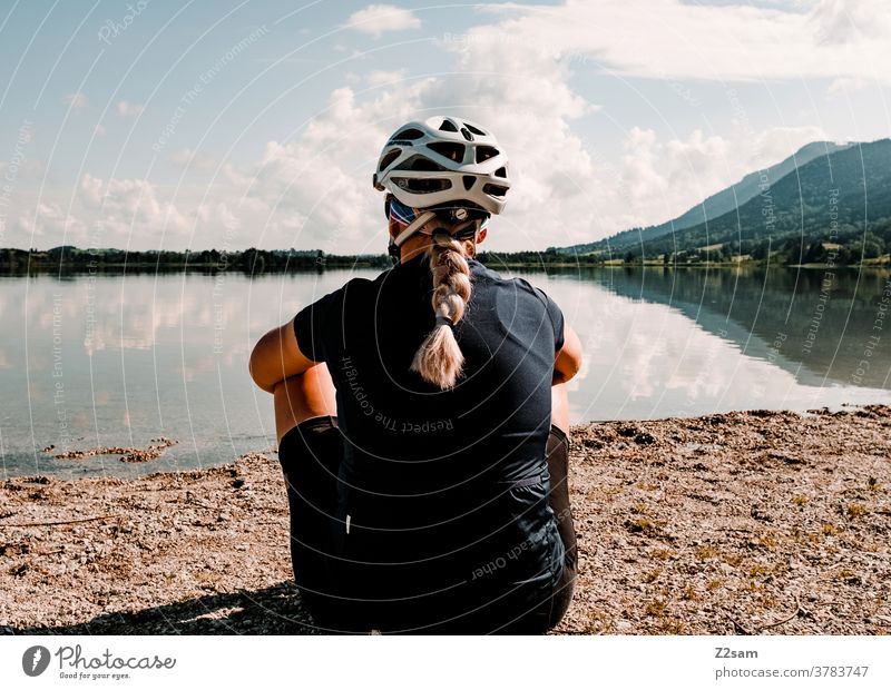 Junger Radfahrer sitzt im Allgäu vor einem See Junge Frau blond Zopf Schutzhelm Radtrikot Rückansicht Erholung Pause Freiheit Wärme Sommer Sonne