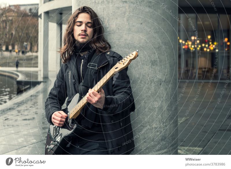 Ein junger Musiker singt im Regierungsviertel Berlin und zupft auf seiner Gitarre Bassgitarre Lied Straßenmusik nachdenklich traurig melancholisch Haare Gebäude