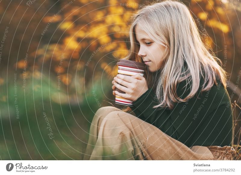 Ein schönes Teenagermädchen mit blonden Haaren sitzt nachdenklich in einem herbstlichen Park, hält eine Thermoskanne mit Tee und wärmt sich die Hände. Herbst