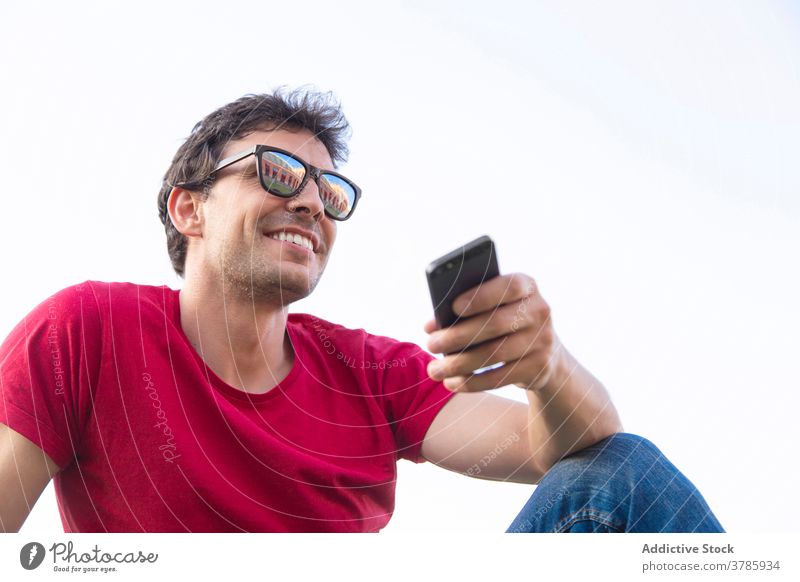 Positiver junger Mann im Park mit Sonnenbrille und Smartphone Fröhlichkeit Lächeln Himmel positiv Lifestyle sich[Akk] entspannen im Freien Person Freude Gras