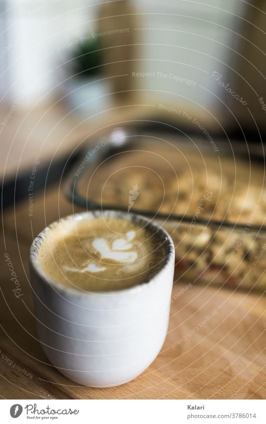 Ein Becher Milchkaffee mit Latte Art Blume auf einem Holztisch mit einem Crumble in einer Auflaufform Kaffee Cappuccino serviert Kuchen Kaffeepause Espresso