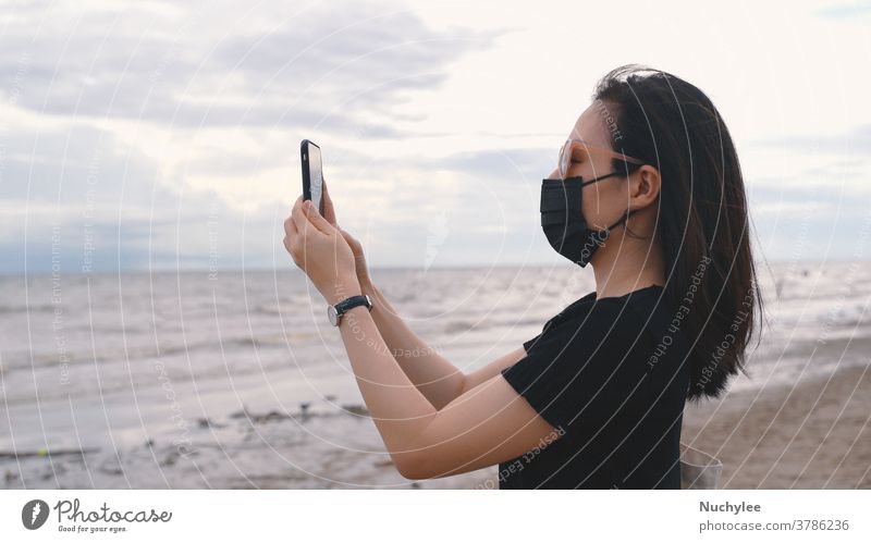 Junge Asiatin mit medizinischer Maske und Smartphone-Fotografie am Strand nach Coronavirus, neuem normalen Lebensstil und Konzept der Reiseblase asiatisch