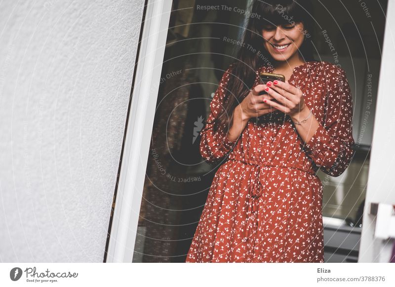 Junge Frau in rotem Kleid mit weißem Muster tippt Lächeln in ihr Smartphone lächelnd benutzend modern zuhause Freude lachen Handy Telefon Mobile