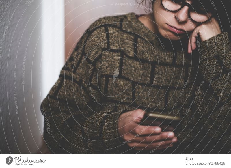 Eine Frau mit Brille guckt gelangweilt in ihr Handy genervt Langeweile Gesicht Traurigkeit Smartphone Mobiltelefon lesen Jugendliche Pullover
