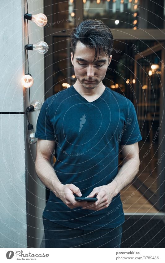 Mann steht vor einem Restaurant im Stadtzentrum und hält ein Smartphone mit Blick auf den Bildschirm Junge Kaukasier Zentrum Großstadt Abend Vorderseite