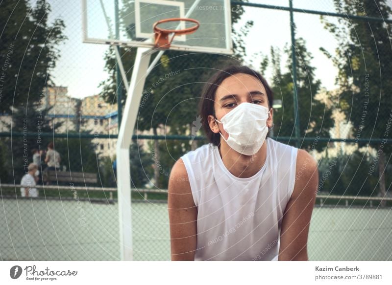 Teenager, der eine Schutzmaske trägt und sich vor einem Basketballkorb ausruht junger Mann männlich Sport im Freien Netz müde vermummt gutaussehend authentisch