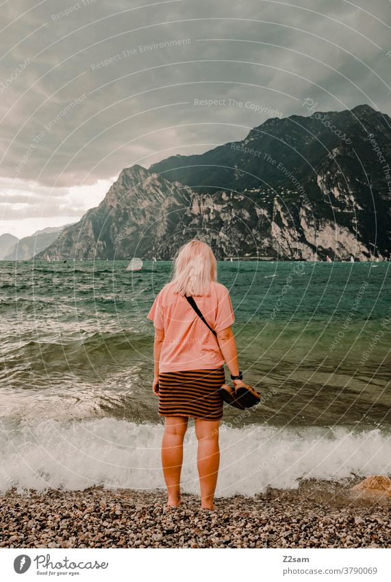 Junge Frau steht am Ufer des Gardasees gardasee norditalien Torbole Urlaub wellen Wasser sehen Gewässer frisch Strang alpenländisch Berge wolken Himmel blau
