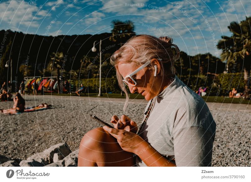 Junge Frau mit Smartphone am Strand von Torbole | Gardasee gardasee Junges Fräulein blond lange haare sonnebrille musik hören Telefon praktisch Strang Natur