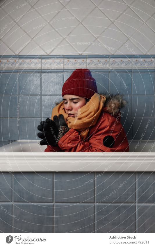 Frau in Oberbekleidung sitzt in der Badewanne in der Nähe der gefliesten Wand kalt Augen geschlossen trendy Bekleidung Fliesen u. Kacheln Konzept heimwärts
