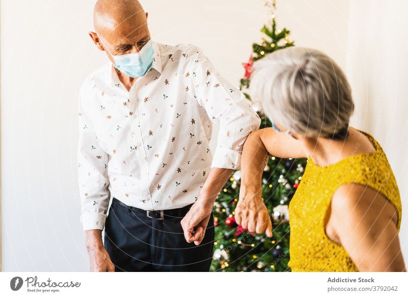 Älteres Paar in Masken stoßen mit Ellbogen in der Nähe von Weihnachtsbaum Begrüßung Weihnachten festlich Vorabend Coronavirus neue Normale Mundschutz salutieren