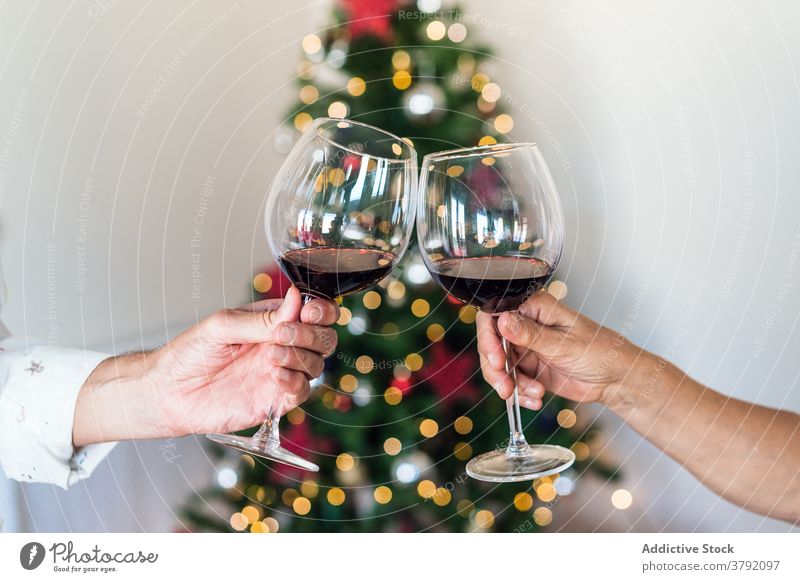 Crop-Paar hebt Gläser mit Rotwein in der Nähe von Weihnachtsbaum Freund Glas jubelt Klirren feiern Weihnachten Baum Neujahr Feiertag festlich glänzend Getränk