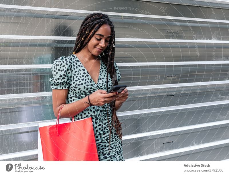 Lächelnde schwarze Frau mit Smartphone auf der Straße Einkaufstasche Tüte konsumgeil benutzend Stil trendy Sommer ethnisch Afroamerikaner Kleid Tasche Großstadt