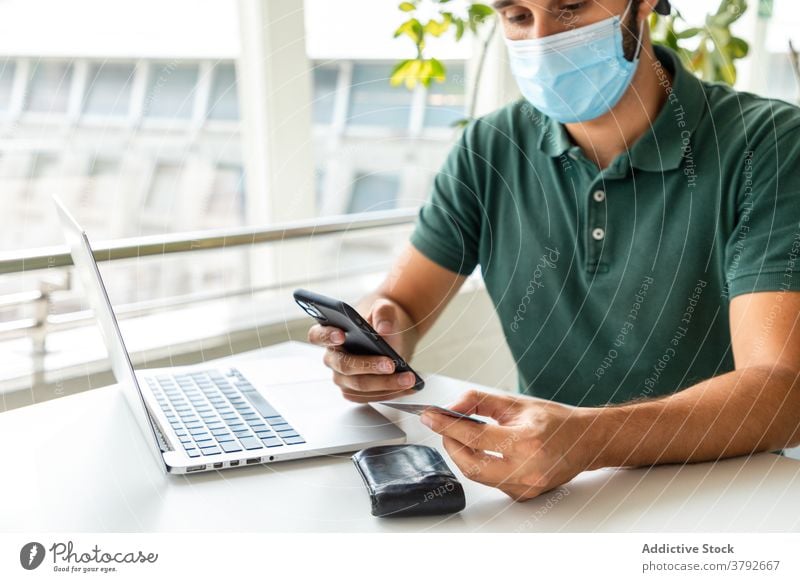 Crop Mann in Maske macht Online-Zahlung Werkstatt online Plastikkarte bezahlen Mundschutz behüten Coronavirus benutzend männlich Smartphone Tisch Windstille