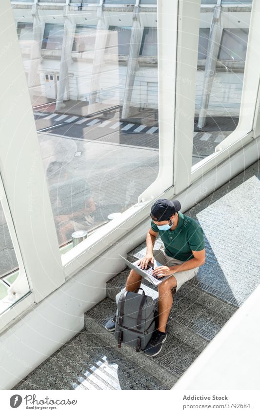 Reisender Mann mit Laptop im Flughafen warten Abheben benutzend Browsen Mundschutz Coronavirus männlich behüten verhindern Netbook Internet sitzen Treppe reisen