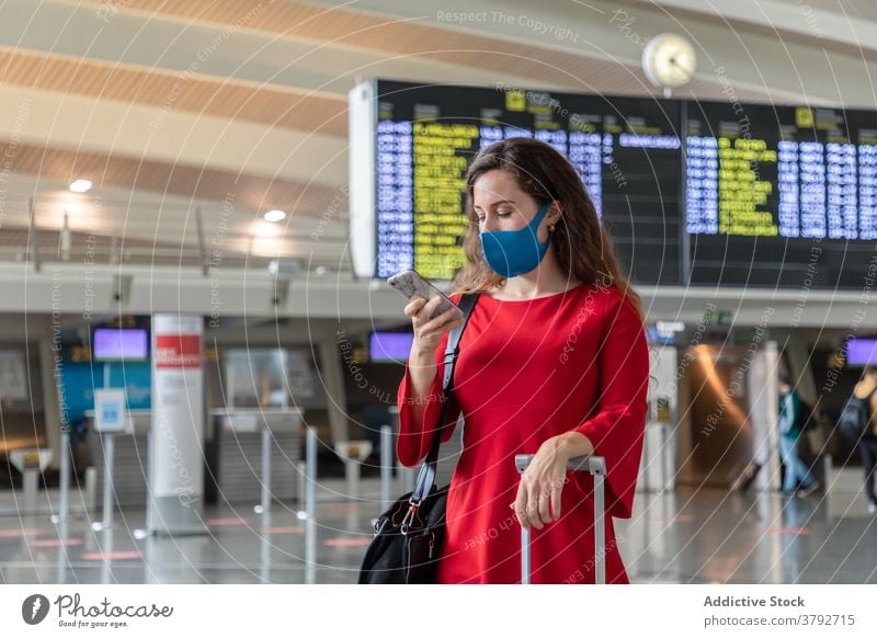 Frau mit Maske benutzt Smartphone im Flughafen Abheben Holzplatte benutzend warten Reisender Mundschutz Coronavirus Koffer Urlaub Gepäck Browsen Funktelefon