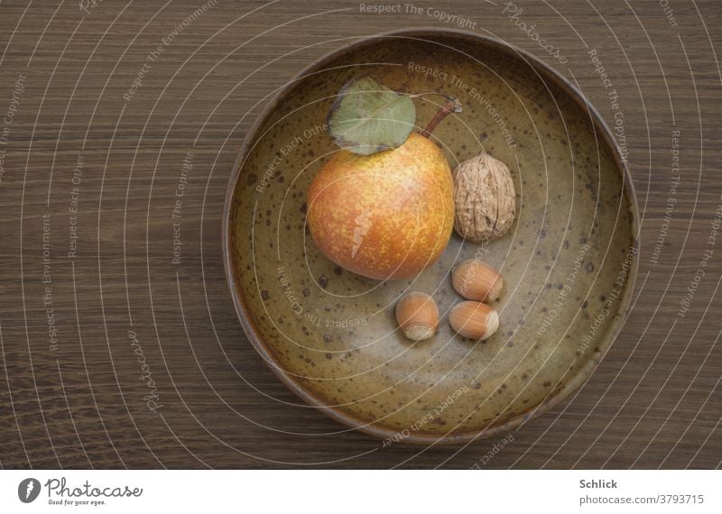 Herbstliche Komposition auf dunklem Eichenholz aus brauner Keramikschale mit Birne Walnuss und drei Haselnüssen Textfreiraum herbstlich Obst monochrom Blatt