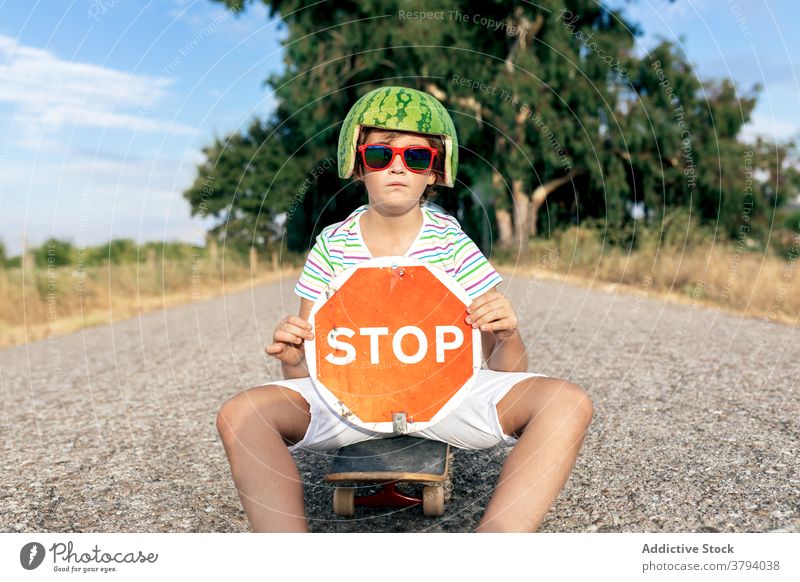 Ernster Junge in dekorativer Kopfbedeckung mit Schild auf der Fahrbahn stoppen verbieten Einschränkung trendy Sonnenbrille Skateboard Straße zeigen Wassermelone