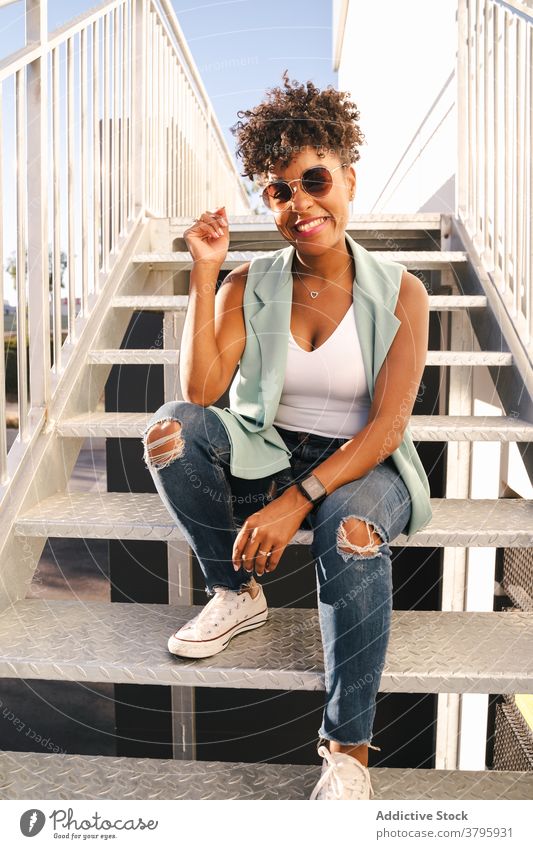 Lächelnde junge schwarze Frau sitzt auf einer Treppe Hipster Stil trendy heiter Glück urban Sonnenbrille modern Afroamerikaner ethnisch Afro-Look Outfit Straße