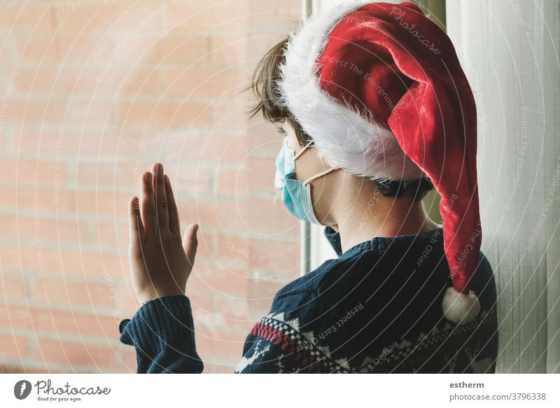 Kind mit medizinischer Maske, das eine Nikolausmütze trägt und an Weihnachten neben einem Fenster auf den Weihnachtsmann wartet Coronavirus medizinische Maske