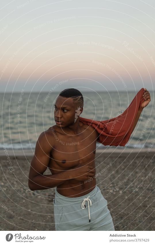 Schwarzer Sportler beim Ausziehen des T-Shirts am Strand stehend Mann muskulös Macho nackter Torso passen stark ohne Hemd Figur brutal männlich sportlich Körper
