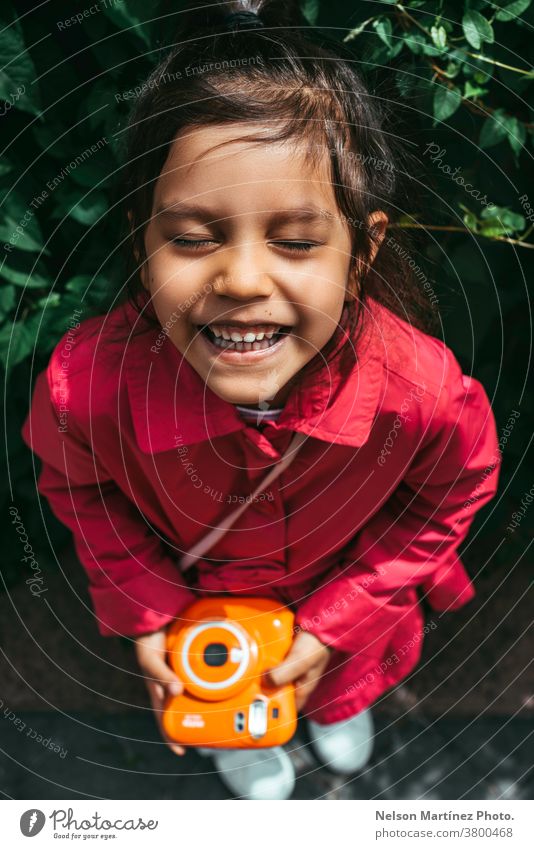 Porträt eines süßen hispanischen Kindes in einer fuchsiafarbenen Jacke. Sie hält eine Polaroidkamera in ihren Händen. Lächeln Fotokamera Fotografie altehrwürdig