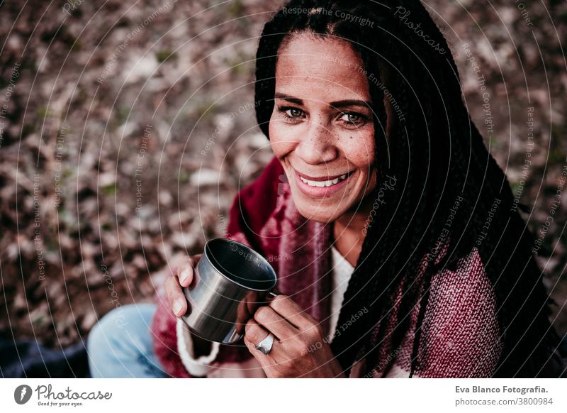 hispanische Frau im mittleren Erwachsenenalter, die im Freien einen Becher Wasser hält. Herbst-Saison Afrofrau Latein Porträt Sonnenuntergang Natur Hut Lächeln