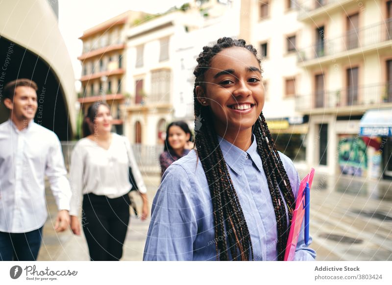 Positiver Schüler mit Ordner hinter multiethnischen Kollegen auf der Straße Lächeln herzlich Geflecht Mappe angenehm freundlich Mitarbeiterin reden Frau