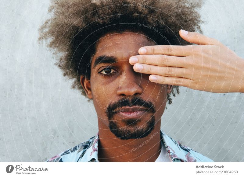 Crop-Partner, der das Auge eines schwarzen Mannes in der Nähe einer Zementwand abdeckt Auge abdecken Freund Fokus stylisch Vorschein Afro-Look Frisur achtsam
