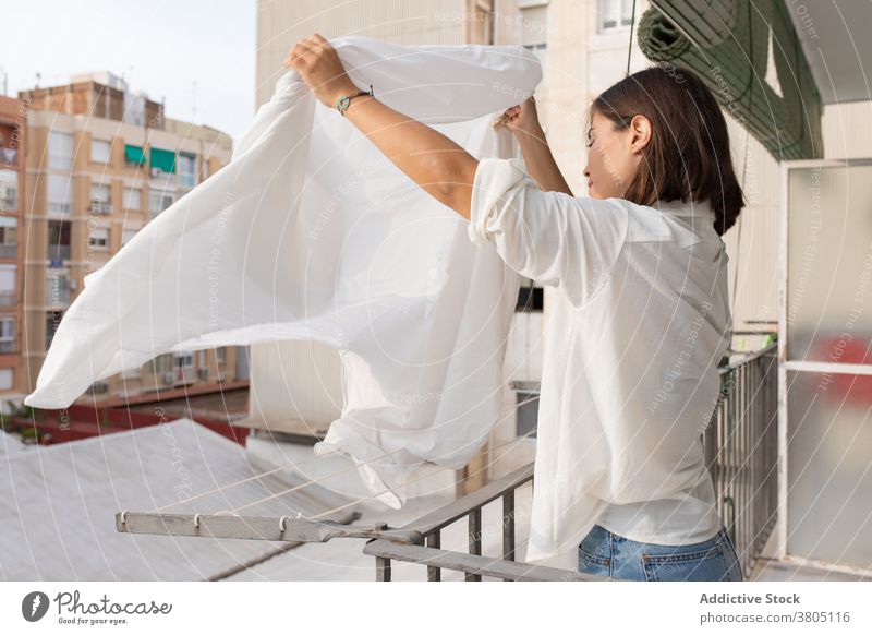 Junge Frau hängt Wäsche auf Balkon Wäscherei Wäscheleine Hausarbeit Waschen heimwärts hängen Bettwäsche Haushalt Hygiene Routine jung weißes Hemd Jeanshose Seil