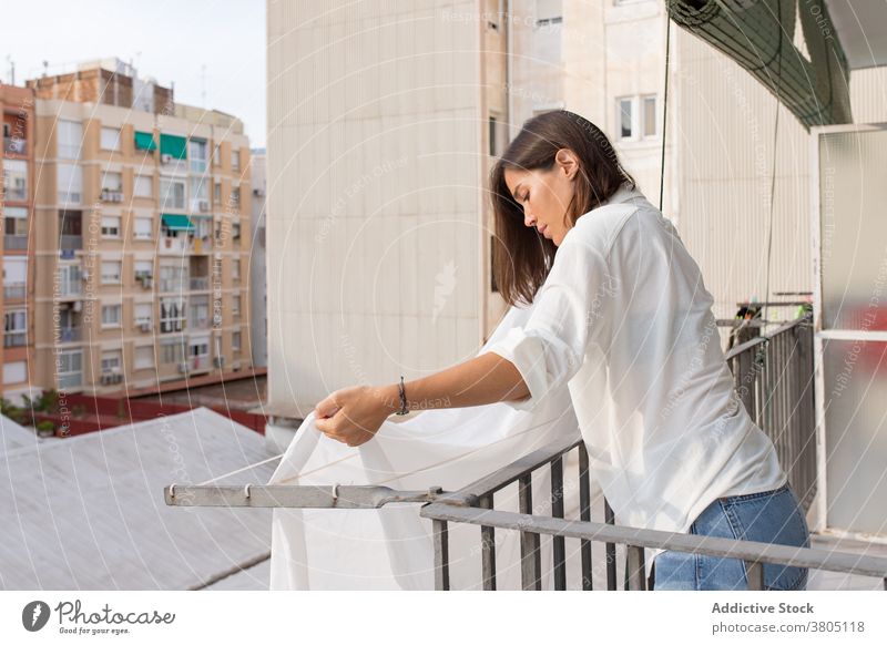 Junge Frau hängt Wäsche auf Balkon Wäscherei Wäscheleine Hausarbeit Waschen heimwärts hängen Bettwäsche Haushalt Hygiene Routine jung weißes Hemd Jeanshose Seil