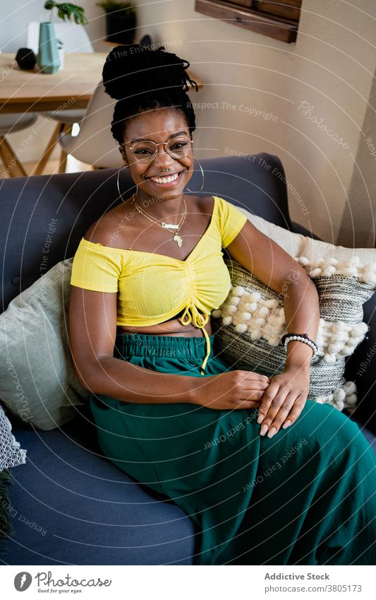 Fröhliche schwarze Frau, die sich im Wohnzimmer ausruht Zahnfarbenes Lächeln Glück zu Hause Stil Mode ruhen Wochenende Freizeit Sofa Afroamerikaner Brille