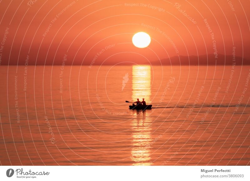 Silhouette von zwei Personen in einem Kanu, die bei Sonnenuntergang an der Spiegelung der Sonne im Meer vorbeifahren. Strand MEER orange Kajak Horizont Browsen