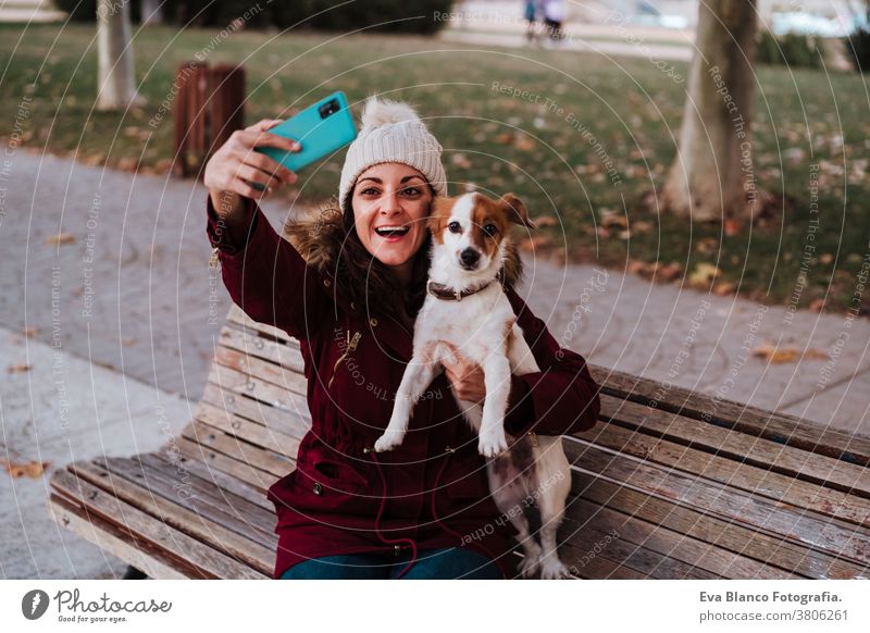 Frau, die auf einer Bank sitzt und in einem Park mit ihrem entzückenden Hund Jack Russell telefoniert. Lebensstil im Freien bezaubernd Herbst Backsteinwand