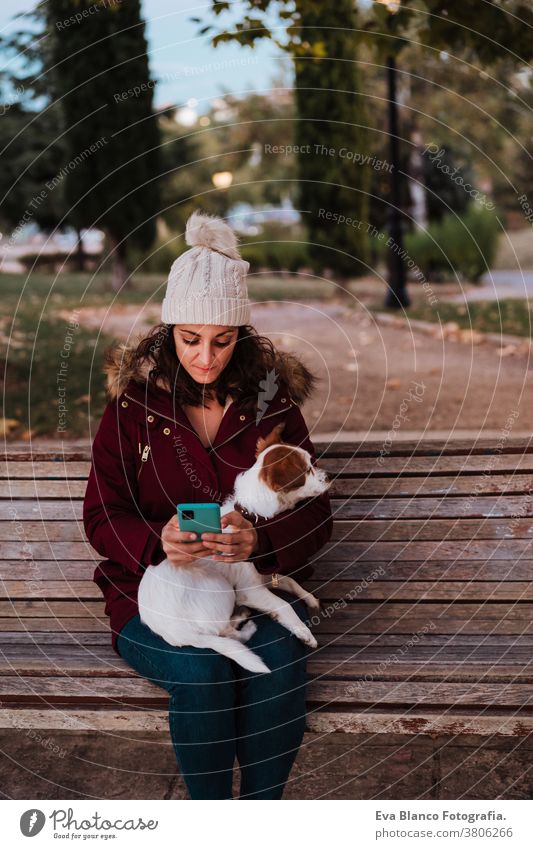 Frau, die auf einer Bank sitzt und in einem Park mit ihrem entzückenden Hund Jack Russell telefoniert. Lebensstil im Freien bezaubernd Herbst Backsteinwand