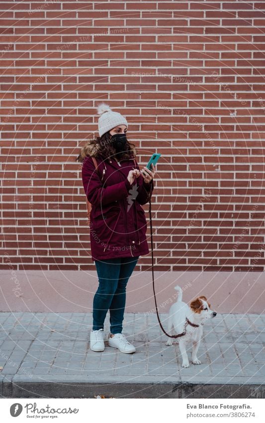 Frau, die eine Schutzmaske trägt, ein Mobiltelefon benutzt und mit ihrem entzückenden Hund Jack Russell durch die Stadt spaziert. Lebensstil im Freien