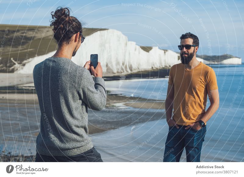 Junger Mann fotografiert mit seinem Smartphone einen Freund, der in der Nähe der Klippe am Meer steht Männer fotografieren Meeresufer Zeit verbringen Feiertag