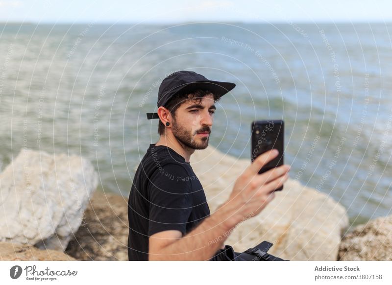 Mann surft im Internet auf dem Smartphone und ruht sich auf Felsbrocken in der Nähe des Meeres aus Selfie MEER Horizont Natur Blauer Himmel benutzend Gerät