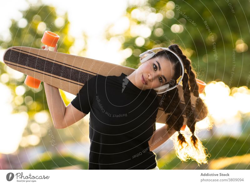 Fröhliche ethnische Dame mit Longboard genießt Musik in Kopfhörer im Park Frau zuhören Skateplatz Glück aktiv heiter trendy Baum Training modern jung