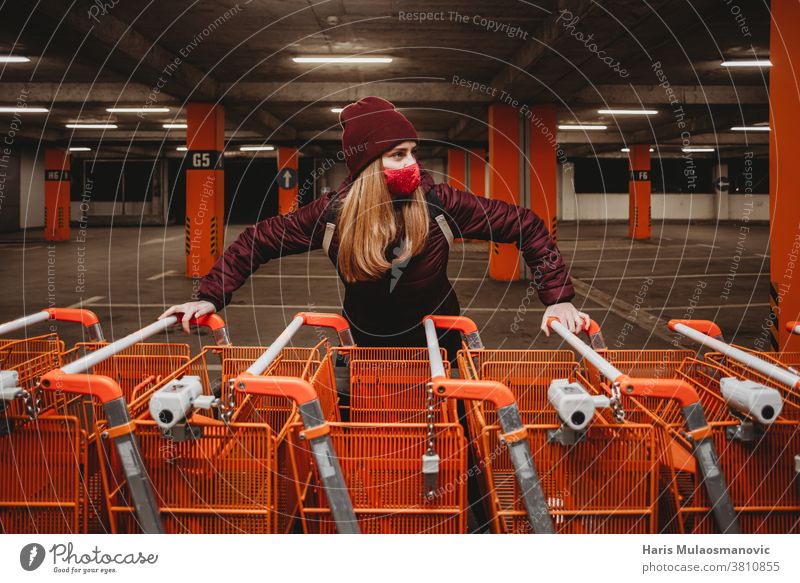 Frau mit Maske, die eine Reihe orangefarbener Einkaufswagen in der Garage hält attraktiv Tasche schön große Einkaufswagen Parkplatz Kaukasier Großstadt Kleidung
