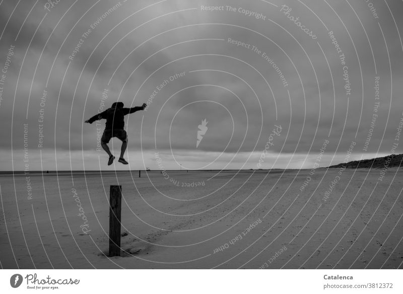 Mann springt von einem Pfosten am Strand. Schwarz-Weiß Tageslicht Nordesee Küste Sand Natur Horizont Landschaft Wasser Meer Himmel Ferien & Urlaub & Reisen