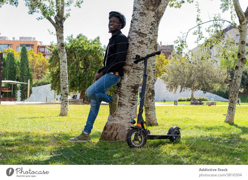Ethnischer Mann in der Nähe von Motorroller im Park Tretroller elektrisch Schutzhelm Großstadt urban Baum Garten behüten männlich ethnisch schwarz
