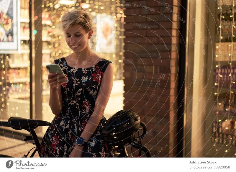 Junge Frau mit Telefon in der Hand sitzt in der Nähe von Geschäft und Fahrrad Kleid Abend Spaziergang Mädchen jung Reiten schön Großstadt Nacht Lichter Gebäude