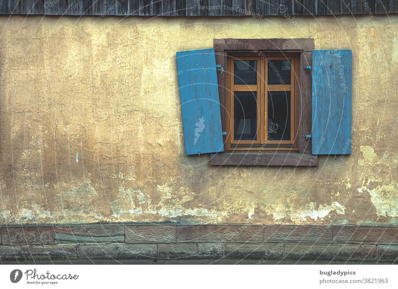 Gelbe/ terrakottafarbene /beige Fassade mit einem braunen Sprossenfenster und blauen / Türkisen Schlagläden. Einer der Schlagläden hängt schief. Fensterladen
