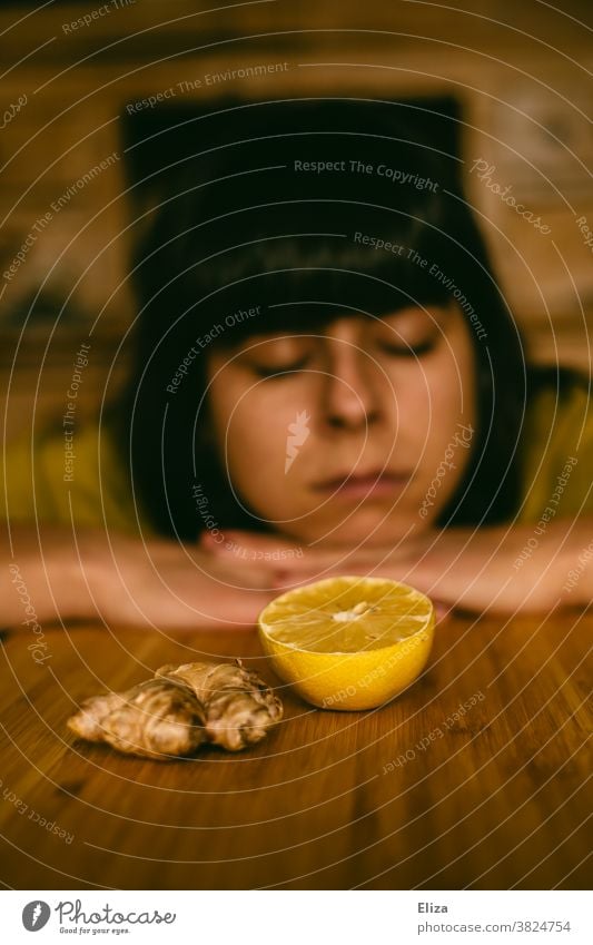 Eine Frau ist müde, schlapp und erkältet und liegt mit dem Kopf auf dem Tisch hinter Ingwer und Zitrone. Erkältung krank Immunsystem Hausmittel