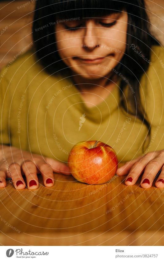 Eine junge frau sitzt am Tisch und blick frustriert auf einen Apfel herab. Diät, gesunde Ernährung. Gesunde Ernährung Vegane Ernährung Frau abnehmen genervt