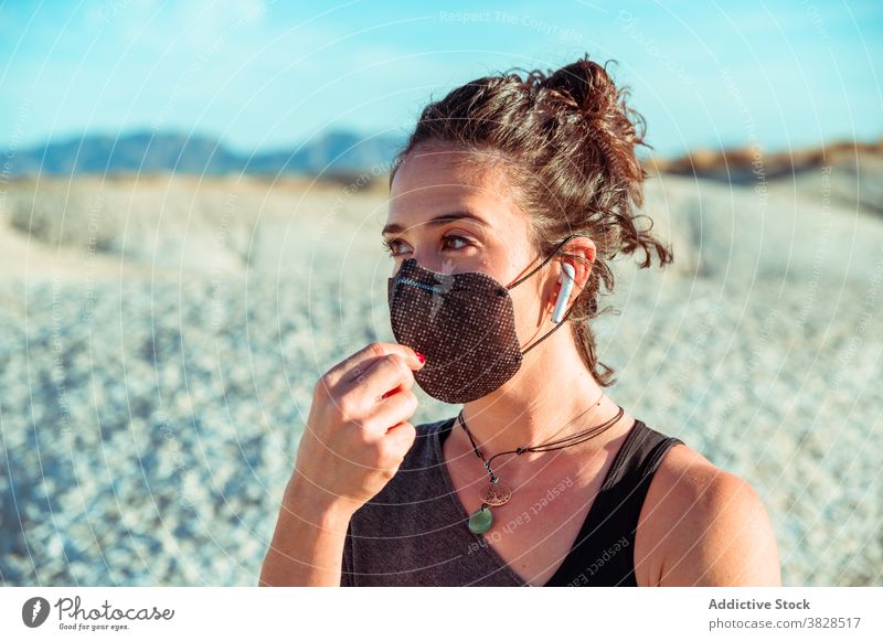 Sportliche Frau mit Maske und Kopfhörern inmitten von trockenem Terrain Sportlerin Mundschutz Ohrstöpsel wüst selbstbewusst zuhören behüten Drahtlos stark