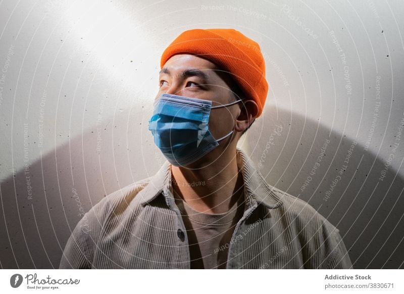Ernster asiatischer Mann mit Gesichtsmaske steht in grauem Korridor Mundschutz Atemschutzgerät Konzentration nachdenklich besinnlich Windstille Deckel Mund