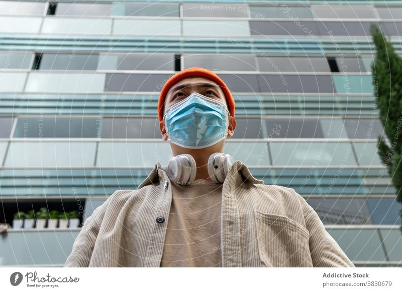 Emotionslos asiatischen Mann in Gesichtsmaske stehen gegen moderne Gebäude emotionslos urban Mundschutz Kondominium Deckel Mund Kopfhörer Atemschutzgerät