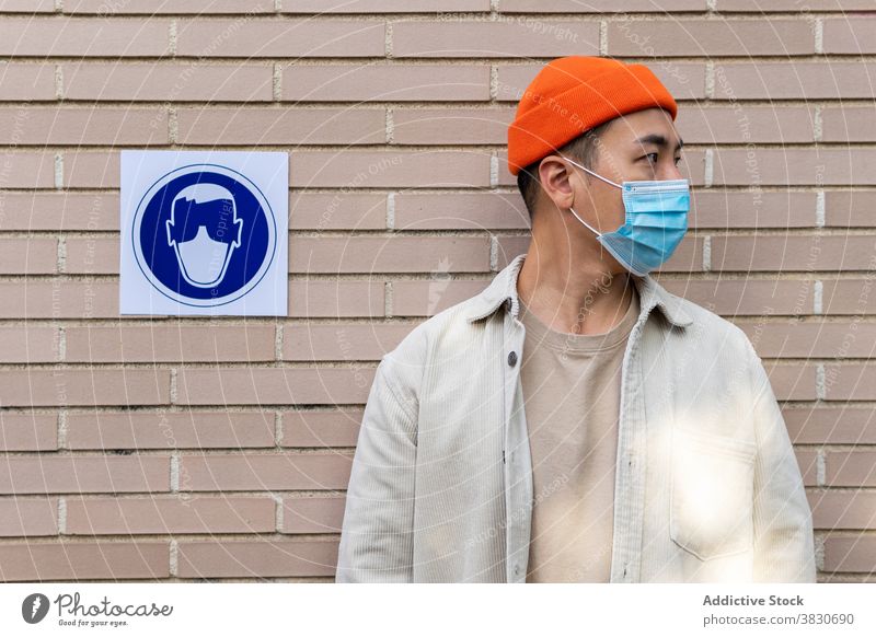 Asiatischer Mann in Maske steht in der Nähe von sing auf Gebäude Wand ernst Mundschutz Zeichen Atemschutzgerät Gehorsam Nachricht Symbol Coronavirus